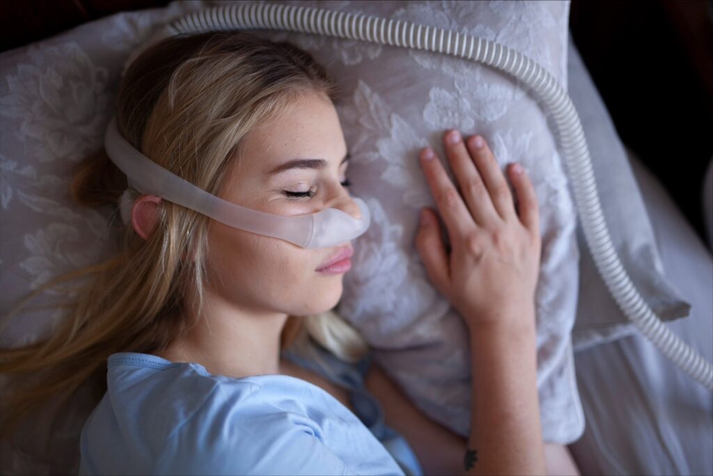 Closeup of young woman with blonde hair laying on her side, resting her head on a pillow as she sleeps with her CPAP obstructive sleep apnea treatment machine on.