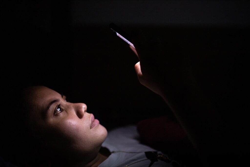 Closeup of young woman laying on her back, in the dark with her phone in her hand, looking right into the bright screen over her phone. The light from the phone illuminates her face, and it looks as though she cannot sleep, so she's just up late on her phone.