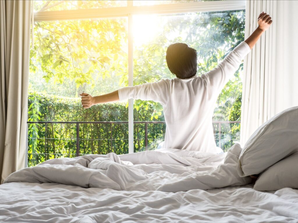 Man sitting on edge of bed, looking out window, stretching with joy after having a good night's sleep.