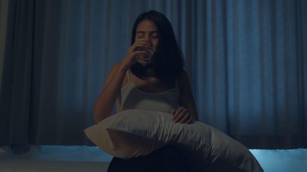 Woman drinking water in the middle of the night with a pillow over her knees, trying hard to get some sleep.