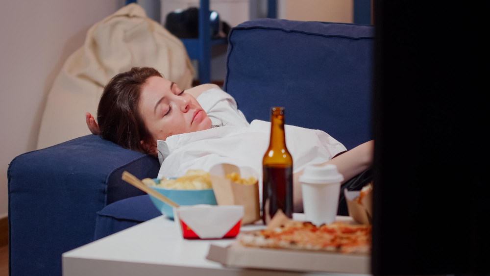 A lady sleeping on the couch with snacks and other eatables in front of a TV in the foreground.