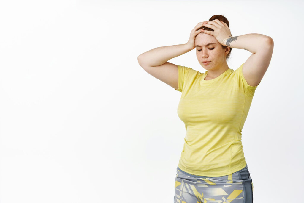 A young plus sized girl grabbing her head with eyes closed, but with workout clothing ready for some exercise but obviously feels tired in the morning.