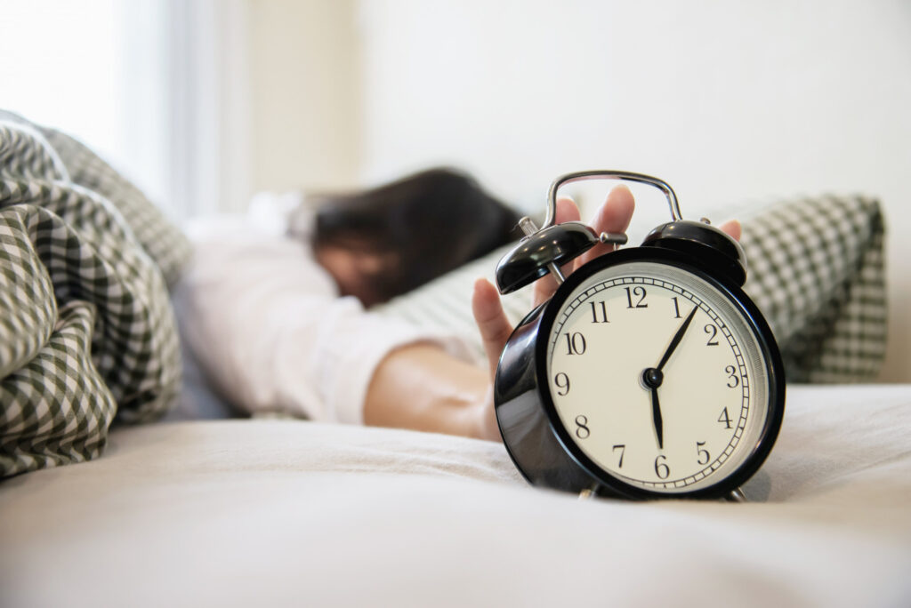 A sleeping person stretching the arm to an alarm clock in the foreground.