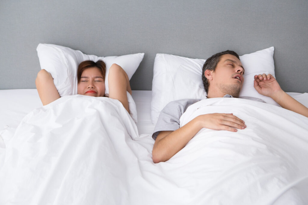 A wife trying to sleep with head between the pillows while the husband snores.