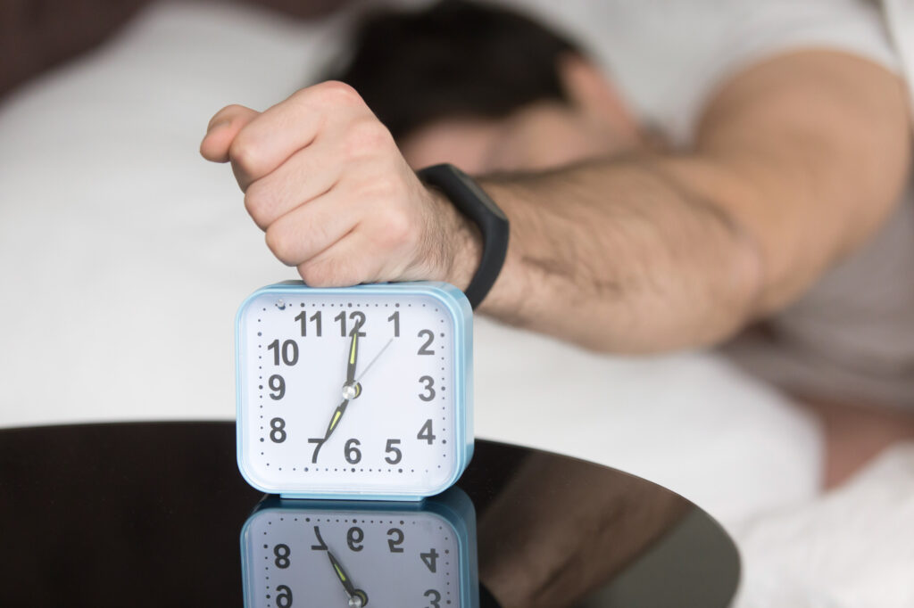 A man is banging an alarm clock while still sleeping