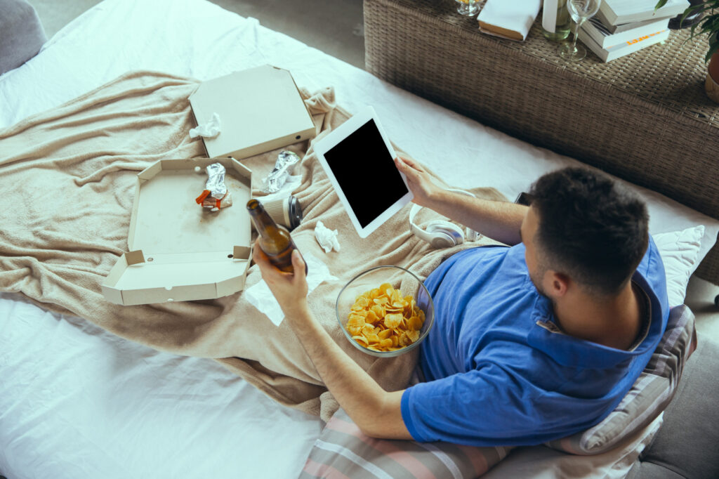 A young man sitting up on a bed with a bowl of chips, a bottle of bear and an empty pizza box