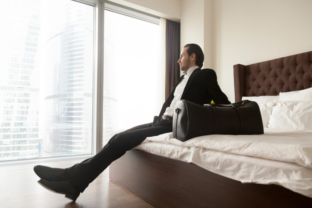 An Affluent traveler and businessman in a hotel room with his bag staring out the window while sitting on the bed