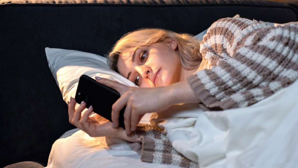 A young lady is on her phone in bed with the lights off and the phone’s light shining across her face