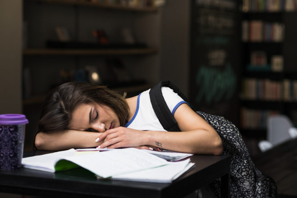 A university student sleeping on a desk at a library with books open in front of her