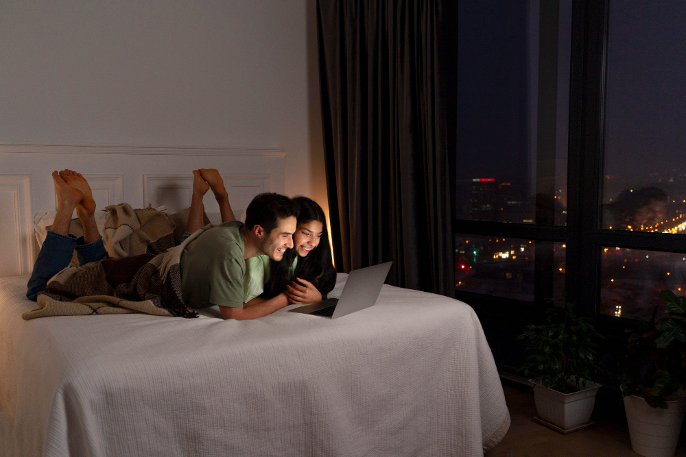 A young couple enjoying a movie night on a loft with dark curtains shading their window that overlooks the city skyline