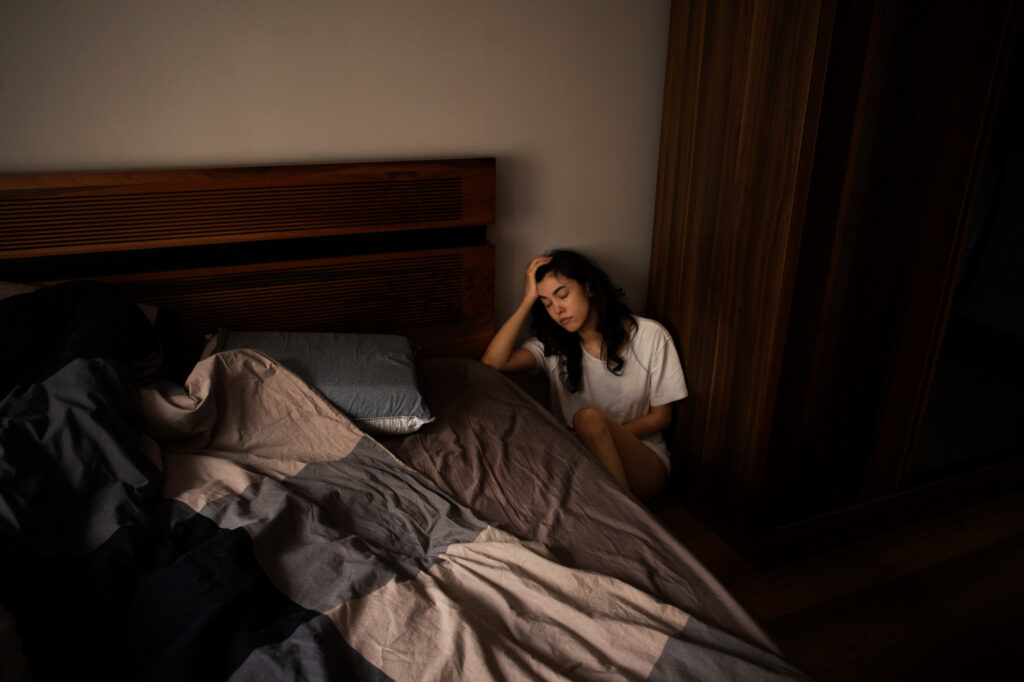 A young woman sitting at the corner of her bed with the sheets piled up trying to find some sleep