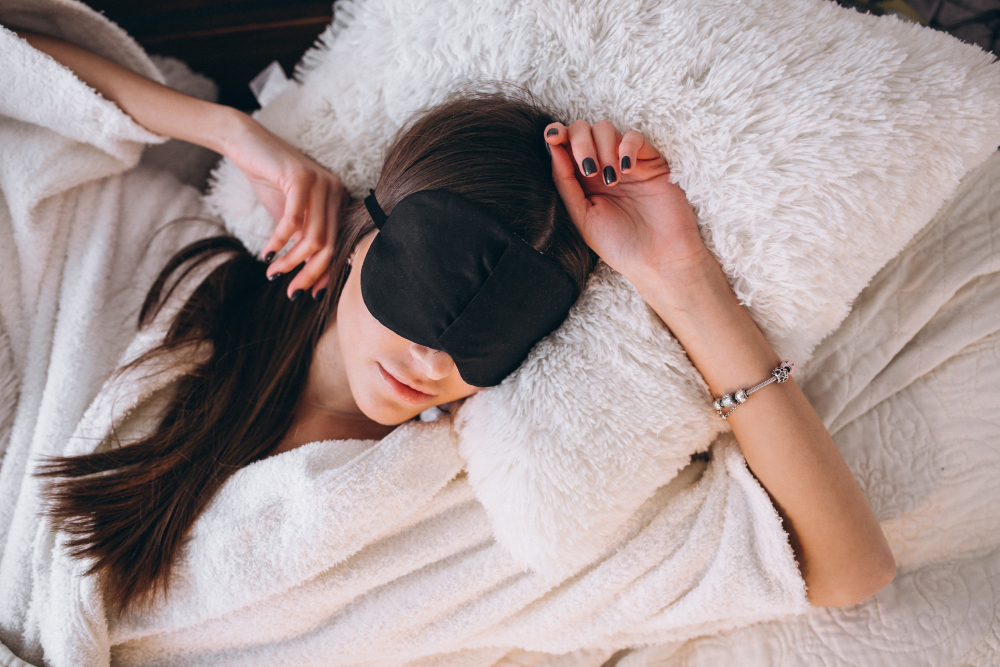A young woman in bed with a bright light shining through taking a power nap using a black eye Mask.